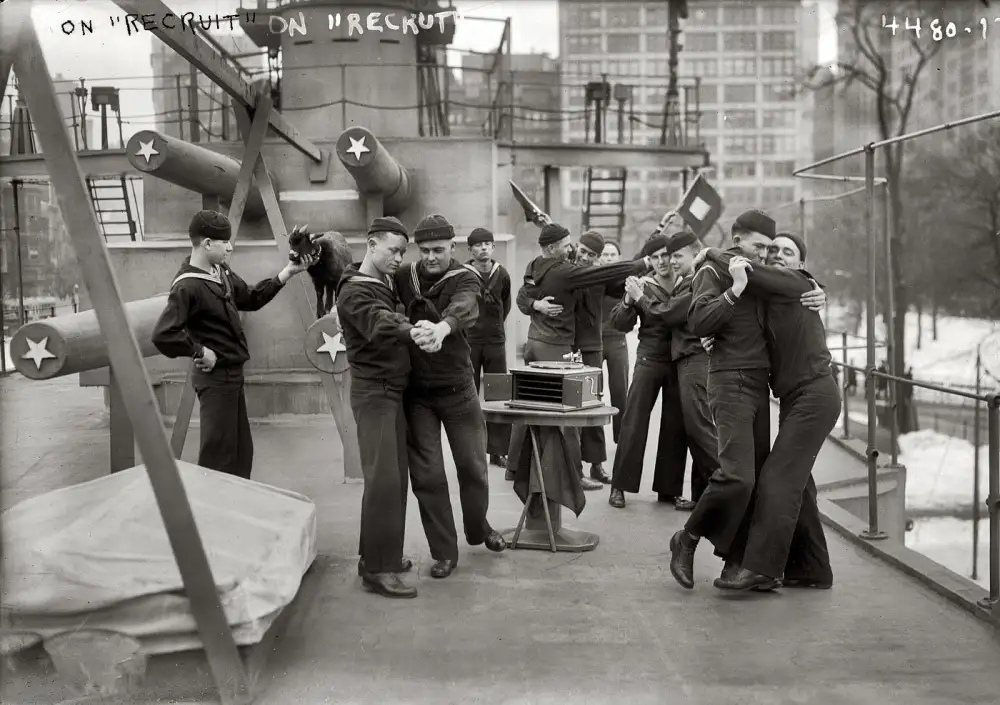 A black-and-white historical photo shows sailors on a ship's deck, some dancing and others interacting near large gun…