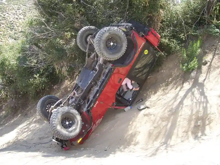 A red Jeep Wrangler is flipped upside down on a sandy, tree-lined hillside, with a person in a pink shirt visible inside the…