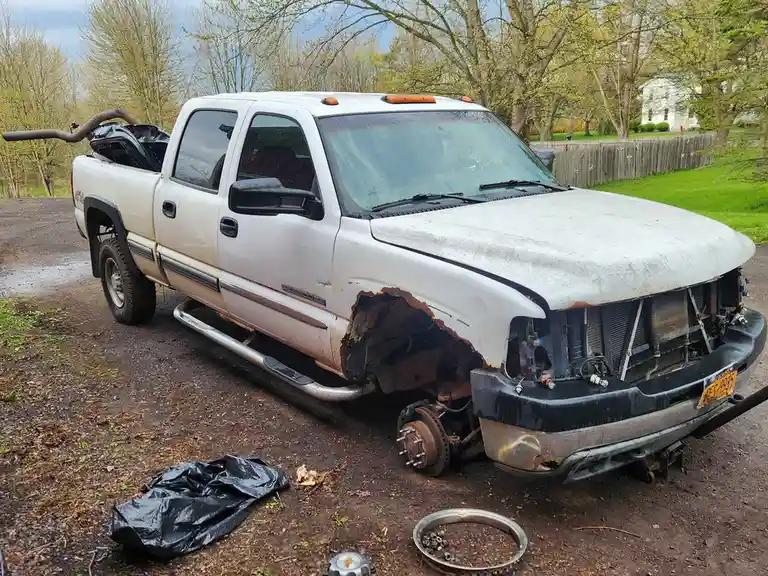 A heavily damaged white GMC Sierra pickup truck is shown with its front end completely stripped, exposing the radiator…