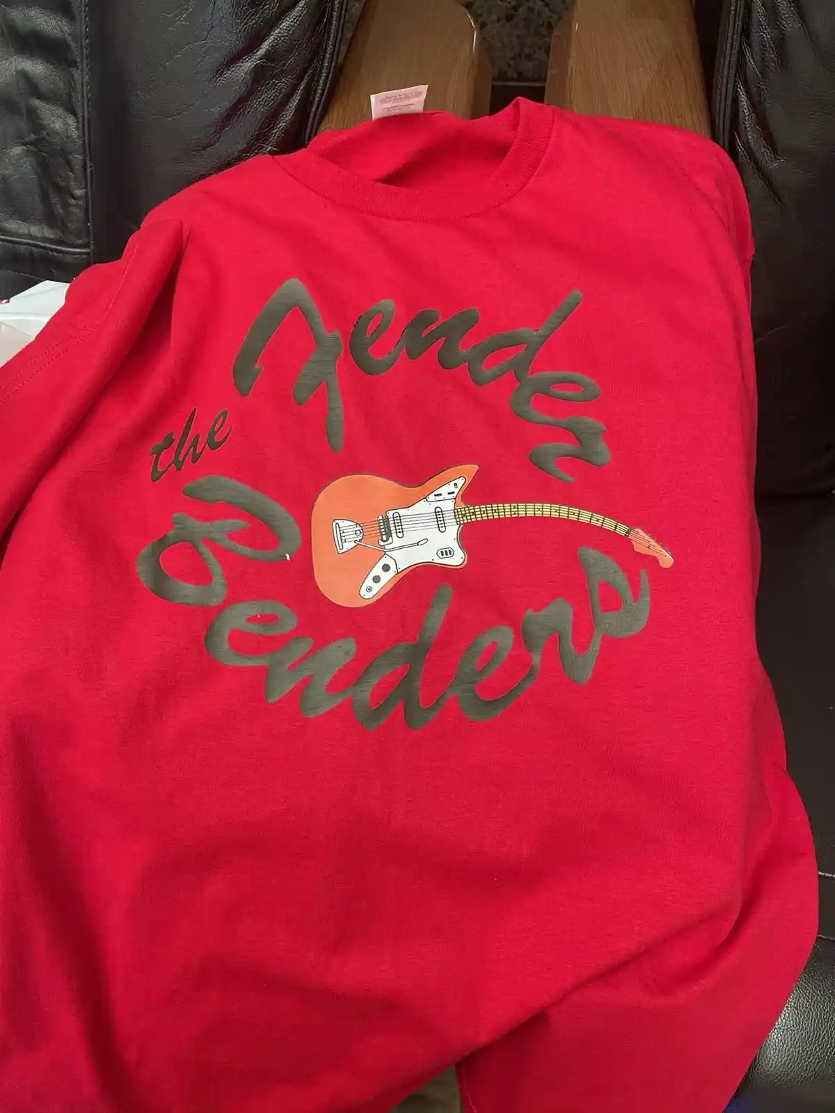 A red t-shirt is laid out on a black leather couch, featuring a graphic of an orange electric guitar with a white pickguard…