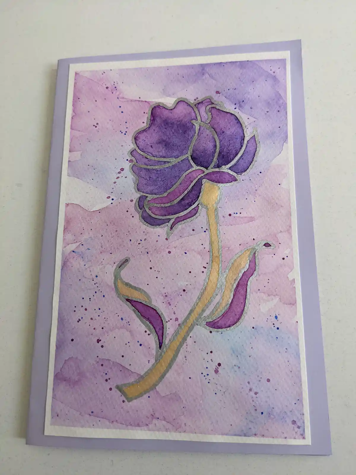 A handmade greeting card features a watercolor illustration of a stylized purple flower with a gold stem and silver foil…
