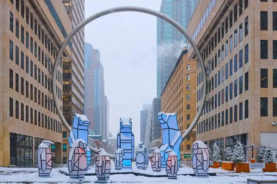 A snowy urban plaza features large, stylized penguin sculptures covered in snow, framed by a giant circular metal arch and…