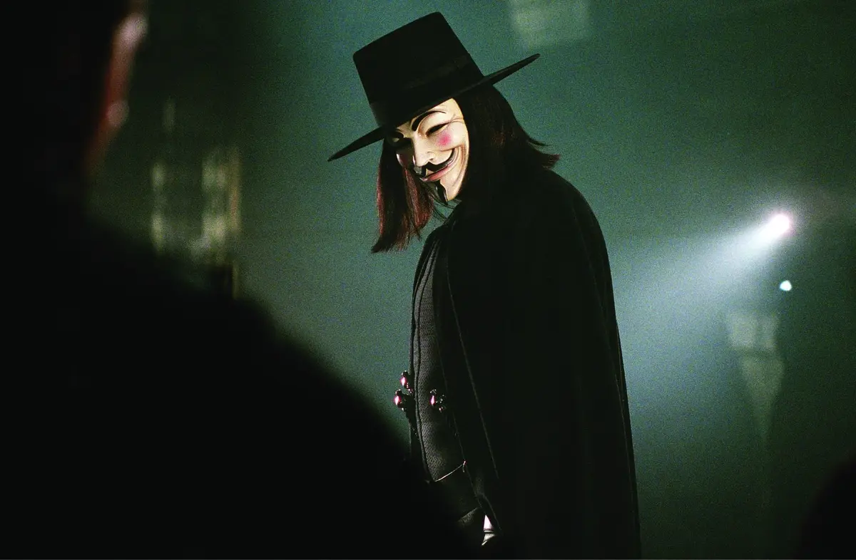 A person wearing a black top hat and a white Guy Fawkes mask with a wide grin, dressed in a black coat, stands in a dimly…