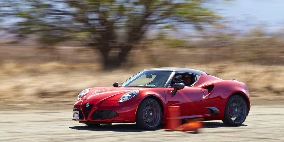 A red Alfa Romeo 4C sports car is captured in motion on a road, with blurred background trees and dry grass indicating high…