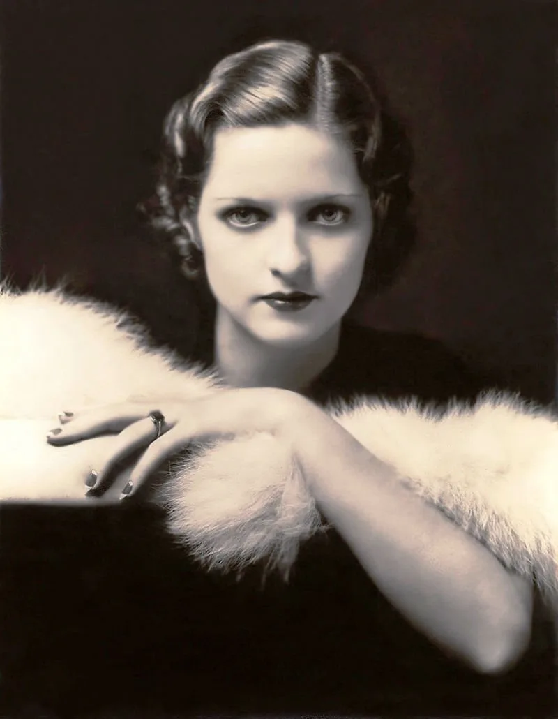 A vintage black-and-white portrait of a woman with styled hair, dark lipstick, and a white fur stole draped over her…