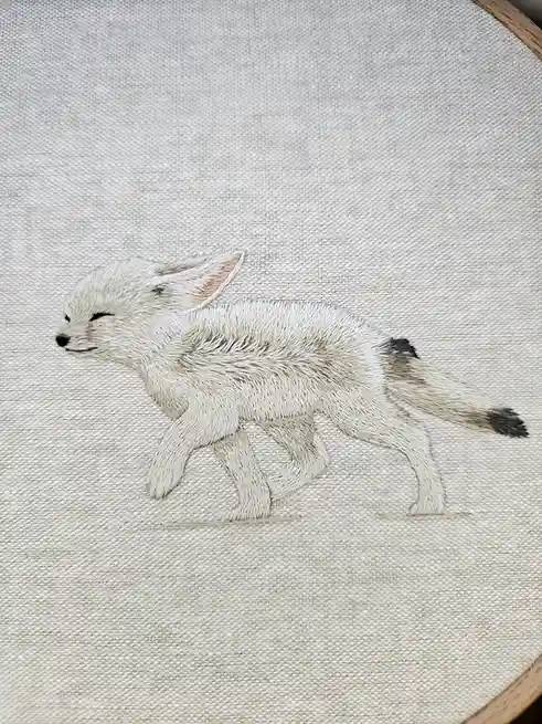 An embroidered fennec fox on light-colored fabric in a hoop, depicted with closed eyes and a serene expression, overlaid…