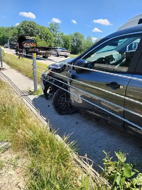 A dark gray car is severely damaged at the front, with its bumper and hood crumpled, parked on the side of a road next to a…