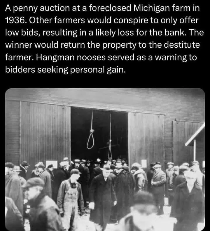 A black-and-white historical photograph shows a crowd of men gathered outside a wooden barn. Suspended from the barn's open…