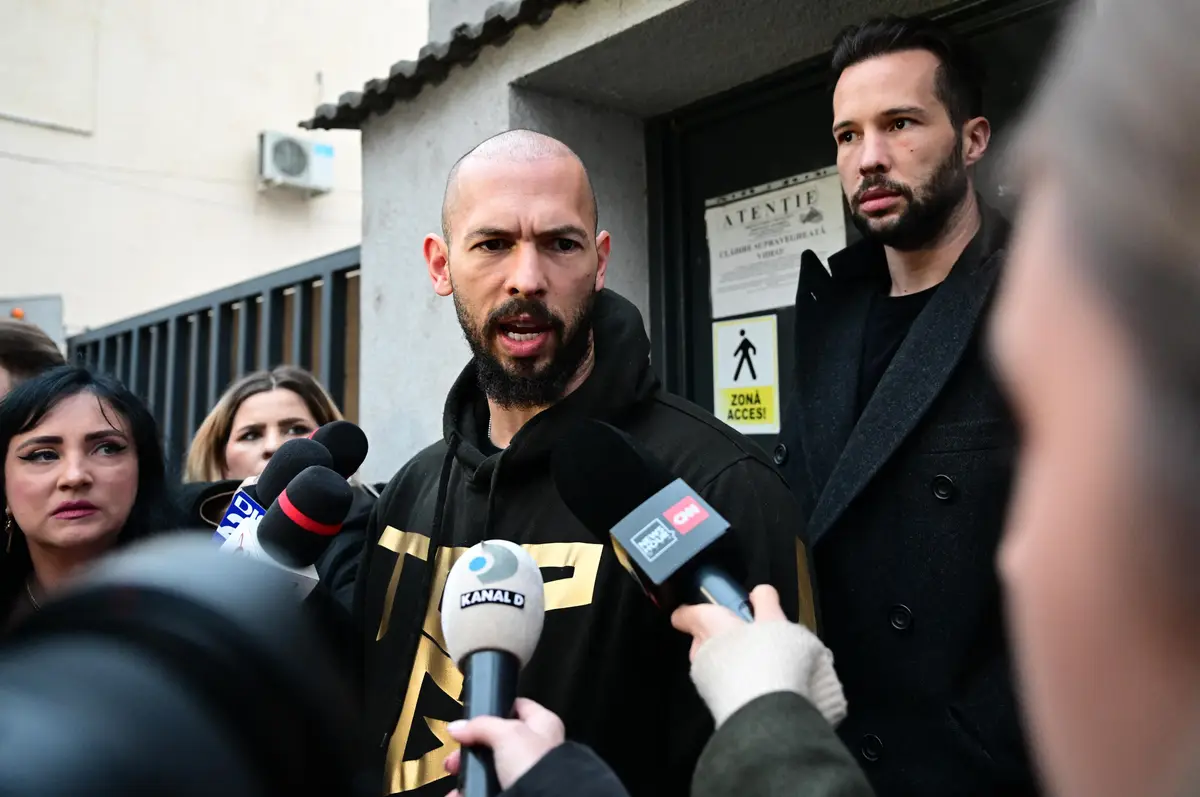 A man in a black hoodie with gold lettering is surrounded by reporters holding microphones from CNN, Kanal D, and other…