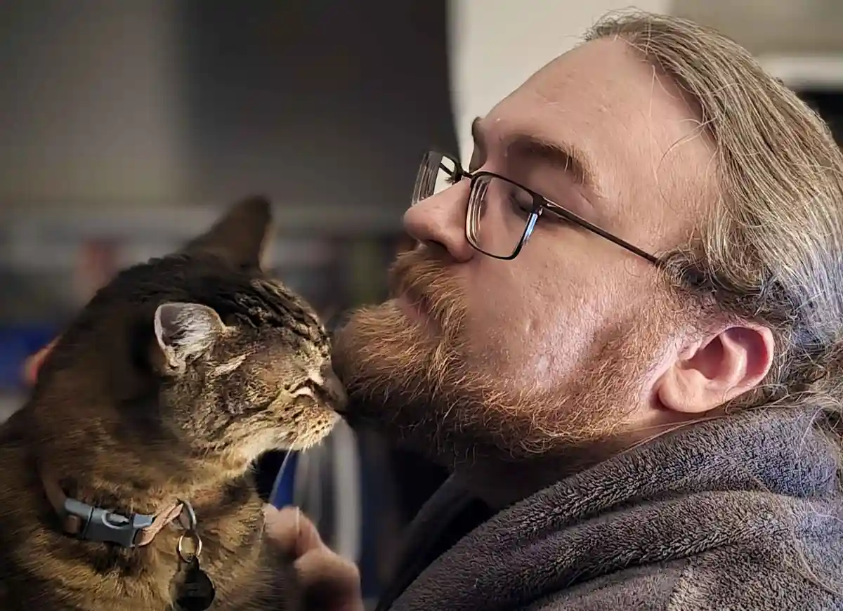 A man with glasses and a beard is shown in profile, gently kissing a tabby cat on the nose. The cat, wearing a collar with a…