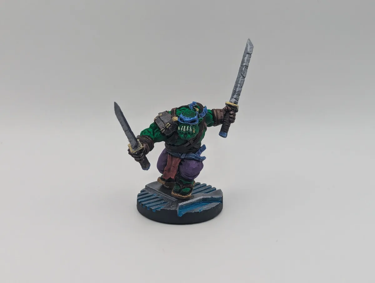 A painted miniature figure of a green-skinned humanoid with a blue mask and purple pants, holding two swords, stands on a…