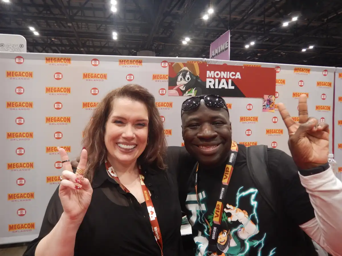 Two smiling people pose for a photo at Megacon Orlando, both making peace signs. Behind them is a branded backdrop with…
