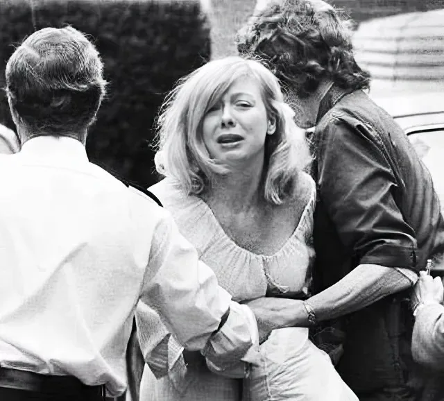 A black-and-white photo shows a distressed woman with blonde hair crying while being held by two men. One man faces away…