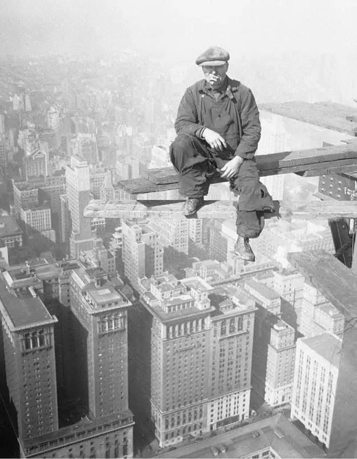 A black-and-white historical photo shows a man in work clothes and a flat cap sitting calmly on a narrow wooden plank…