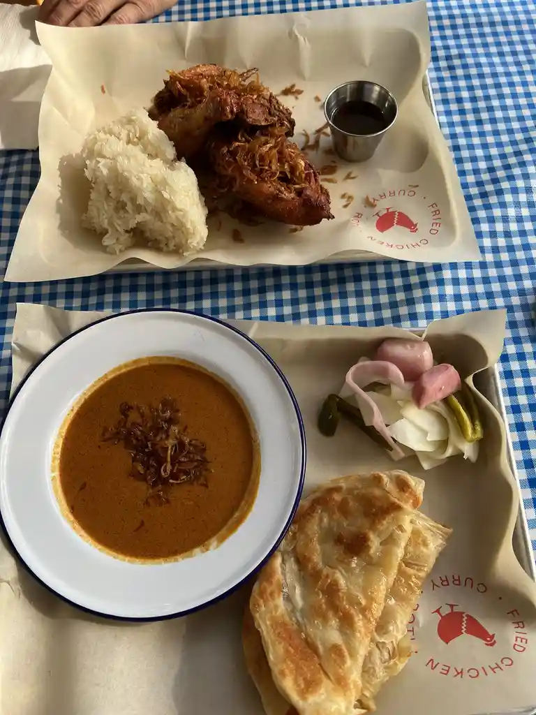 Two trays of food on a blue-and-white checkered tablecloth. One tray holds fried chicken with rice and a dipping sauce. The…