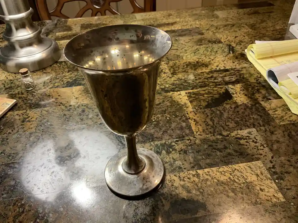 A silver, stemmed wine glass with a wide bowl sits on a speckled granite countertop, covered in condensation droplets. The…