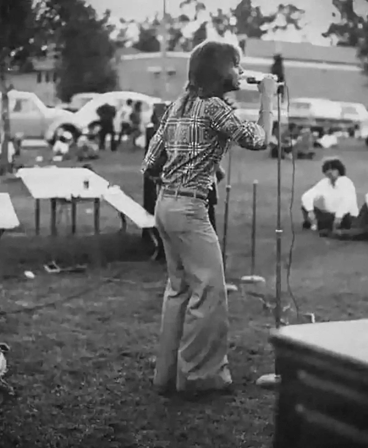 A black-and-white photo shows a person in a plaid shirt and wide-leg pants singing into a microphone outdoors, with people…
