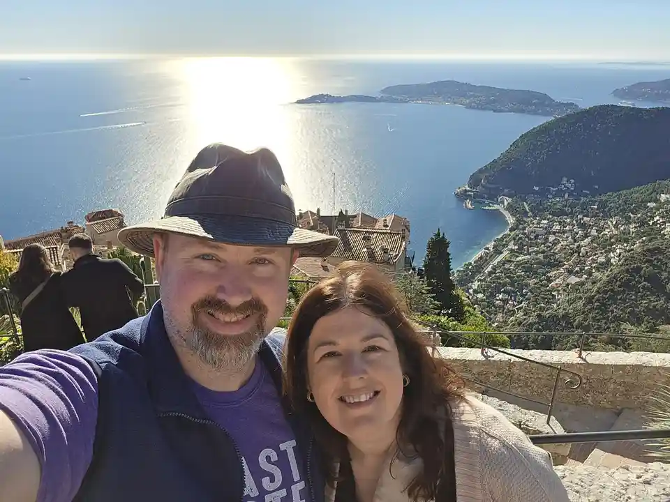 A smiling man and woman take a selfie on a scenic overlook with a stunning view of the ocean, islands, and coastal town…