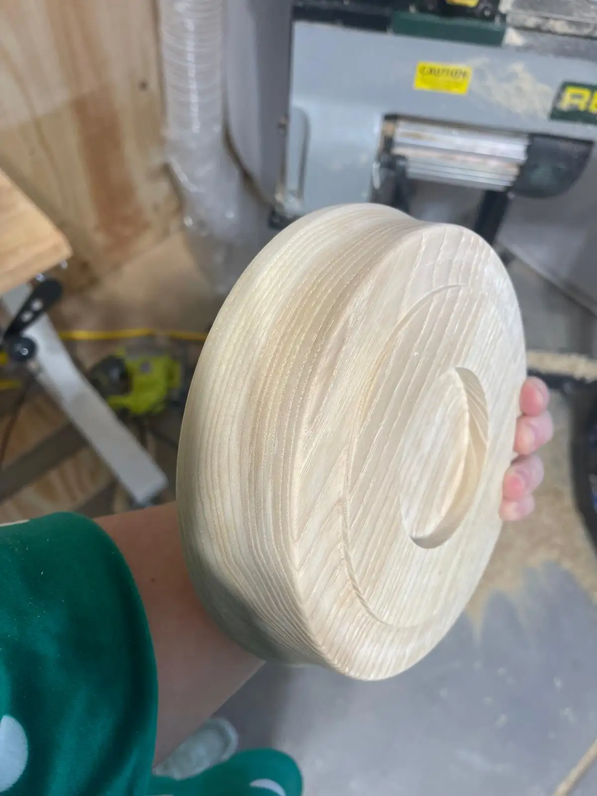A person holds up a freshly crafted wooden bowl in a workshop, showcasing its smooth, light-colored grain and a small handle…