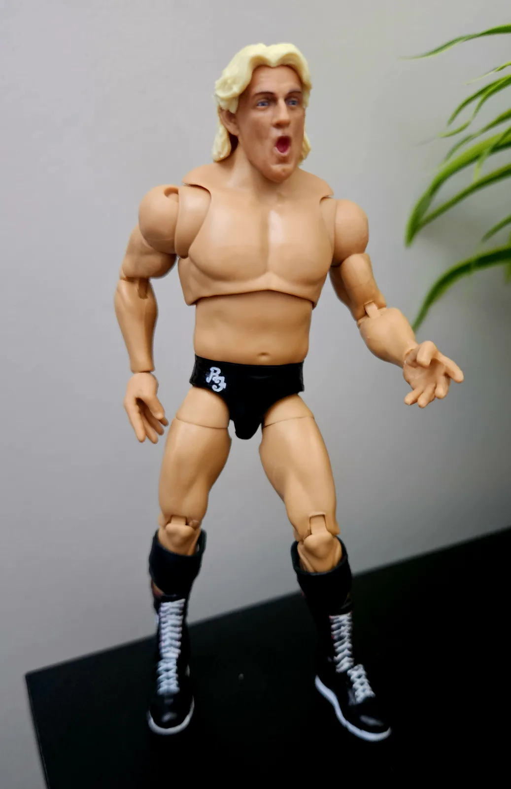A muscular action figure with blonde hair, wearing black wrestling trunks with a white logo and black boots with white…
