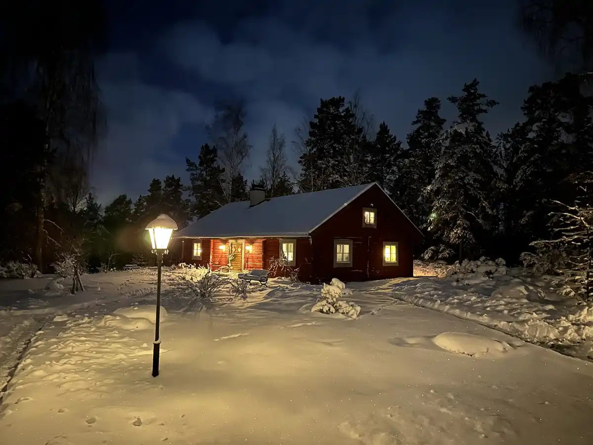 A cozy red wooden cabin is illuminated at night, surrounded by deep snow and snow-covered trees. A lit streetlamp stands in…