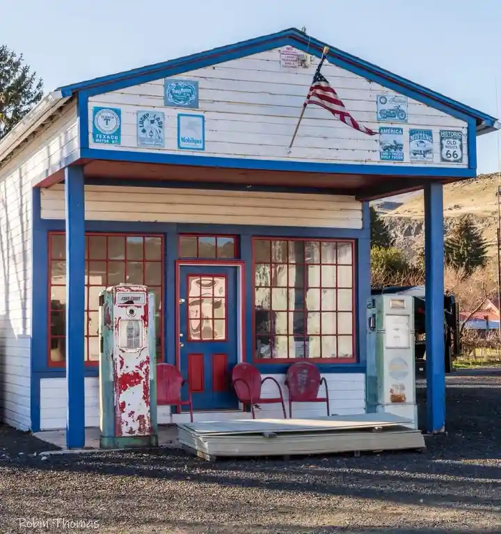 A vintage roadside gas station with blue and white siding, red-trimmed windows, and two weathered gas pumps. An American…