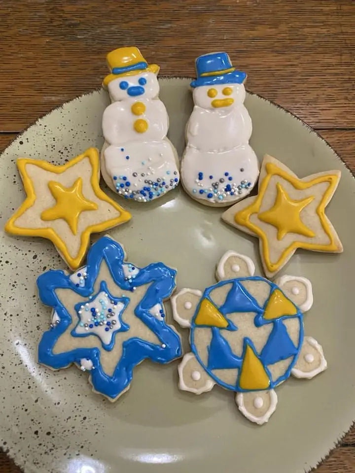 A plate of six decorated sugar cookies sits on a wooden table. Two snowman-shaped cookies with blue and yellow hats and…