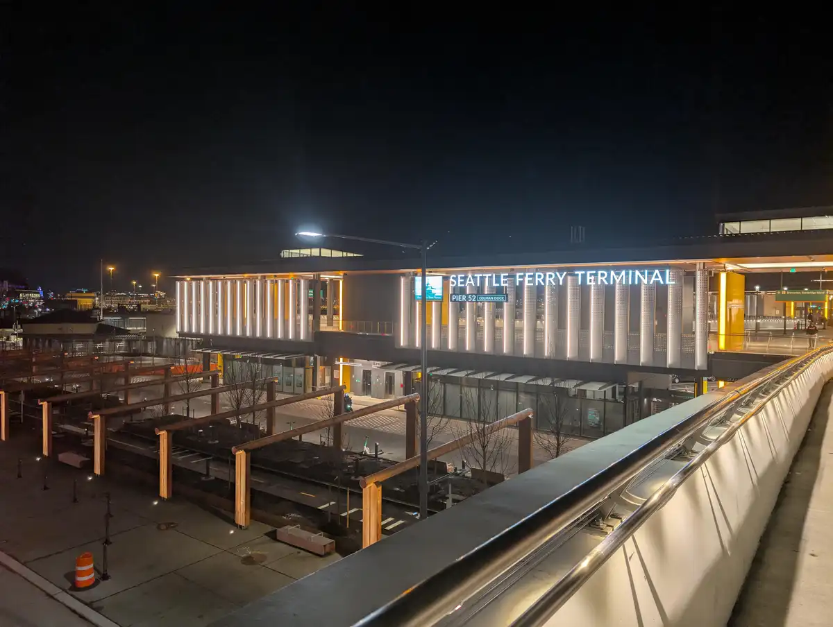 A nighttime photo of the modern Seattle Ferry Terminal building, illuminated with bright lights, showing its large sign…