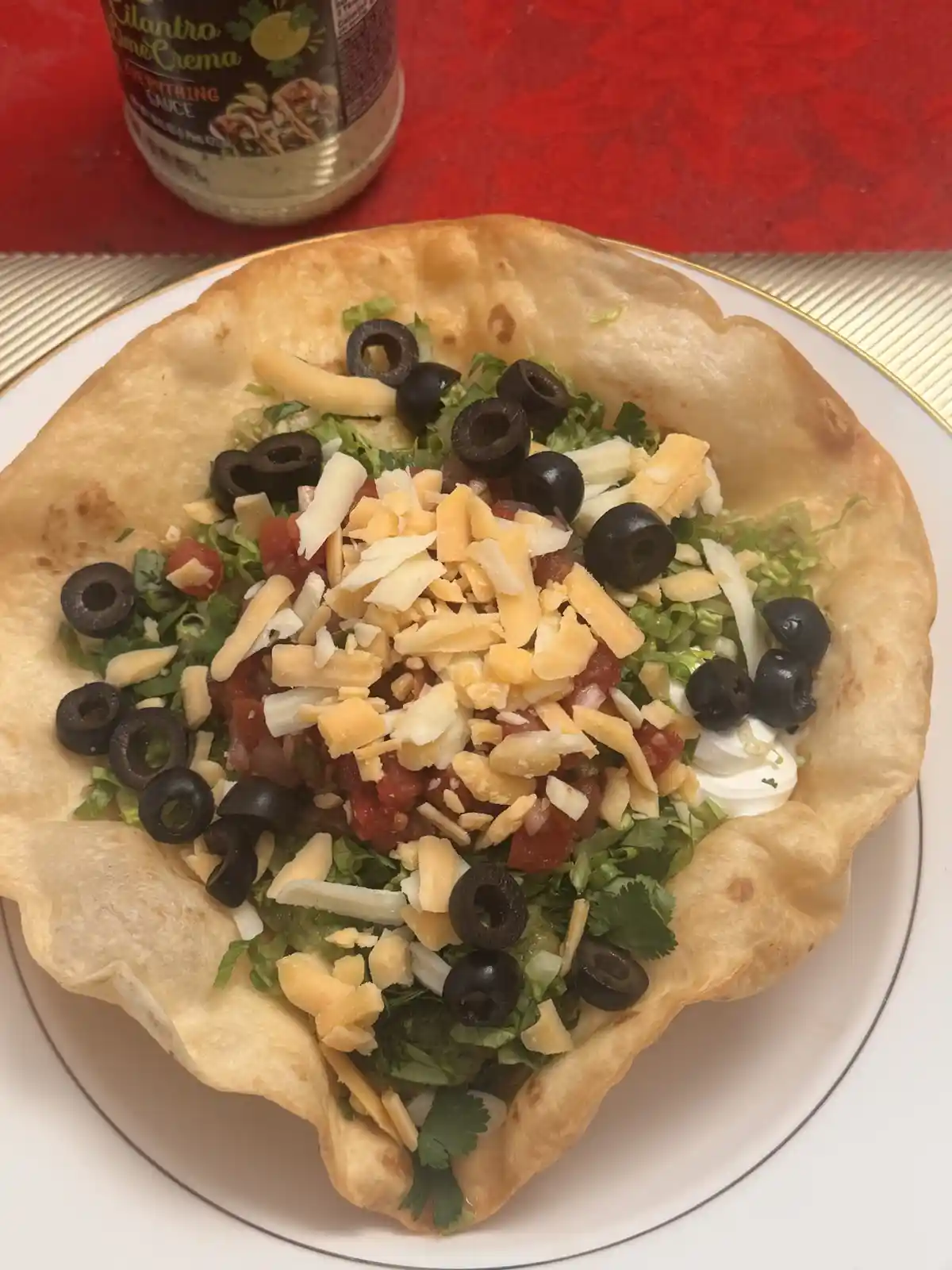 A close-up photo shows a crispy tortilla bowl filled with lettuce, black olives, shredded cheese, diced tomatoes, and a…