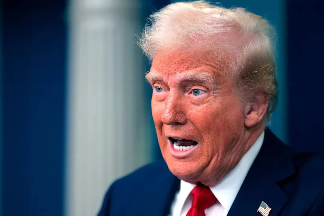 A close-up photo of Donald Trump, wearing a dark suit, white shirt, and red tie with an American flag pin, looking to his…