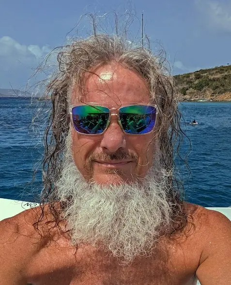 A man with long, wet, gray hair and a full white beard wears reflective sunglasses while on a boat. The background shows…
