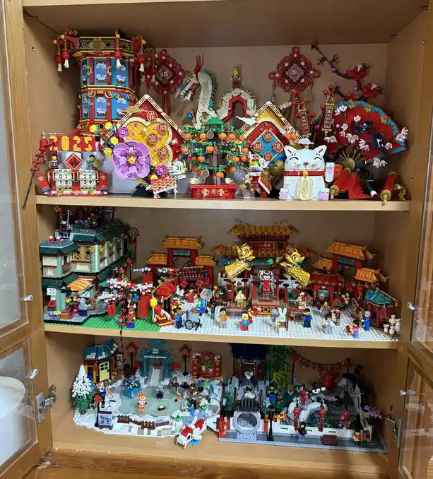 A wooden display cabinet with three shelves filled with intricate Lego models themed around Chinese New Year and traditional…