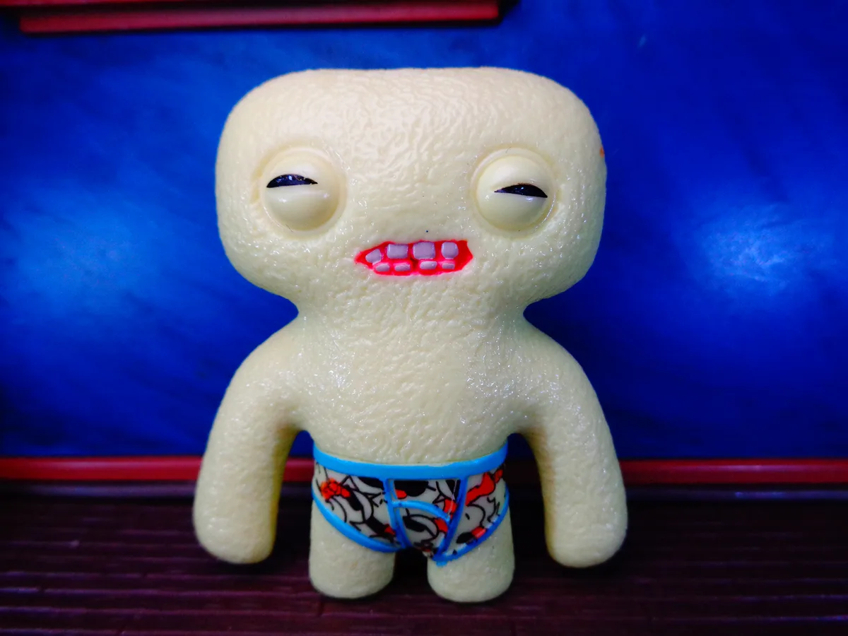 A pale, textured, Uglydoll-like toy with large, half-closed eyes and a red, toothy grin stands against a blue background. It…