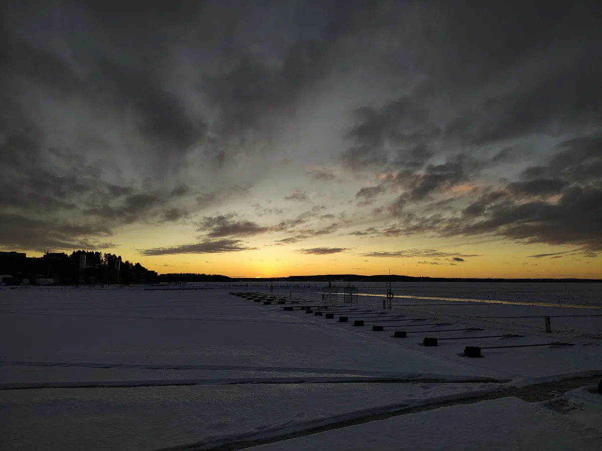 A wide shot of a frozen lake at sunset, with dark clouds overhead and a golden horizon. The ice is marked with rows of black…