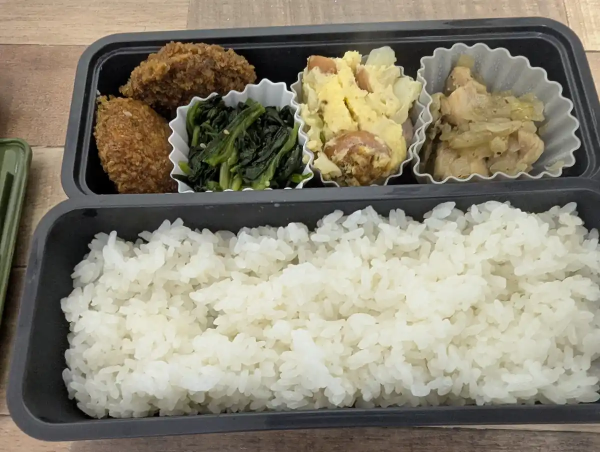 a black bento box contains a large portion of white rice in the bottom compartment and four smaller compartments with fried…