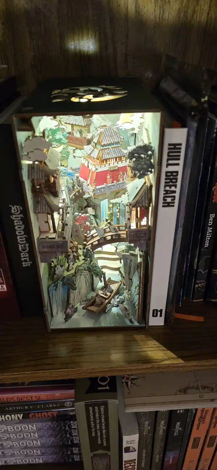 A lit, intricate 3D paper model of a traditional Chinese temple village sits on a bookshelf, flanked by books including one…