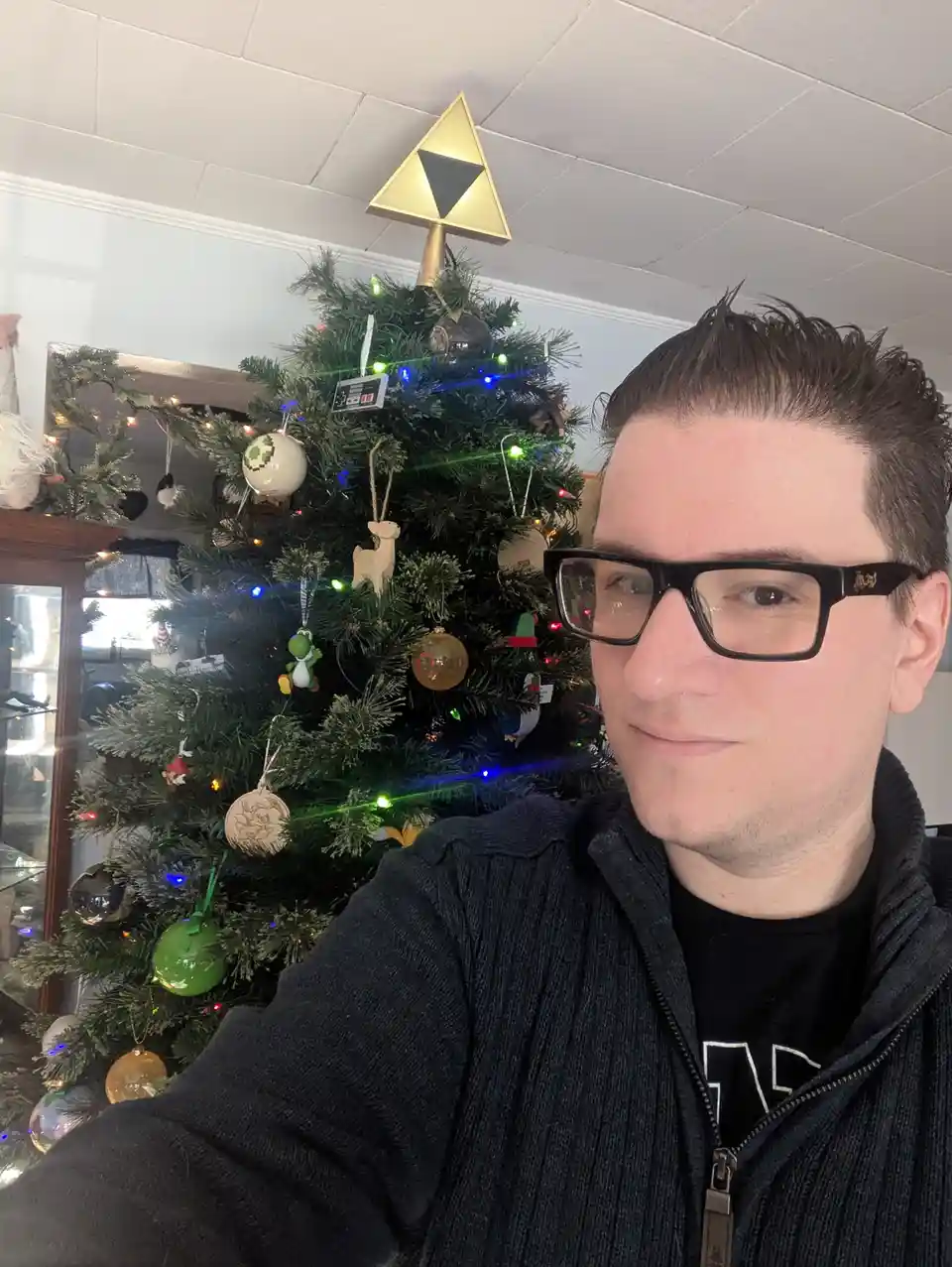 A man with glasses takes a selfie in front of a decorated Christmas tree adorned with video game-themed ornaments, including…