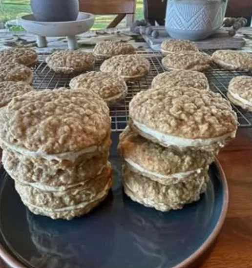 A photo of oatmeal cookies, some stacked in towers on a dark blue plate, others cooling on a wire rack. The cookies have a…