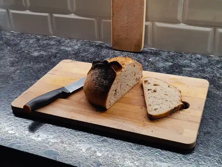 A loaf of bread with one side severely burnt sits on a wooden cutting board next to a single slice and a serrated knife…