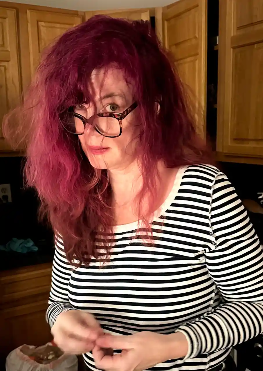 A person with long, messy magenta hair and glasses, wearing a black-and-white striped shirt, looks at the camera with a…