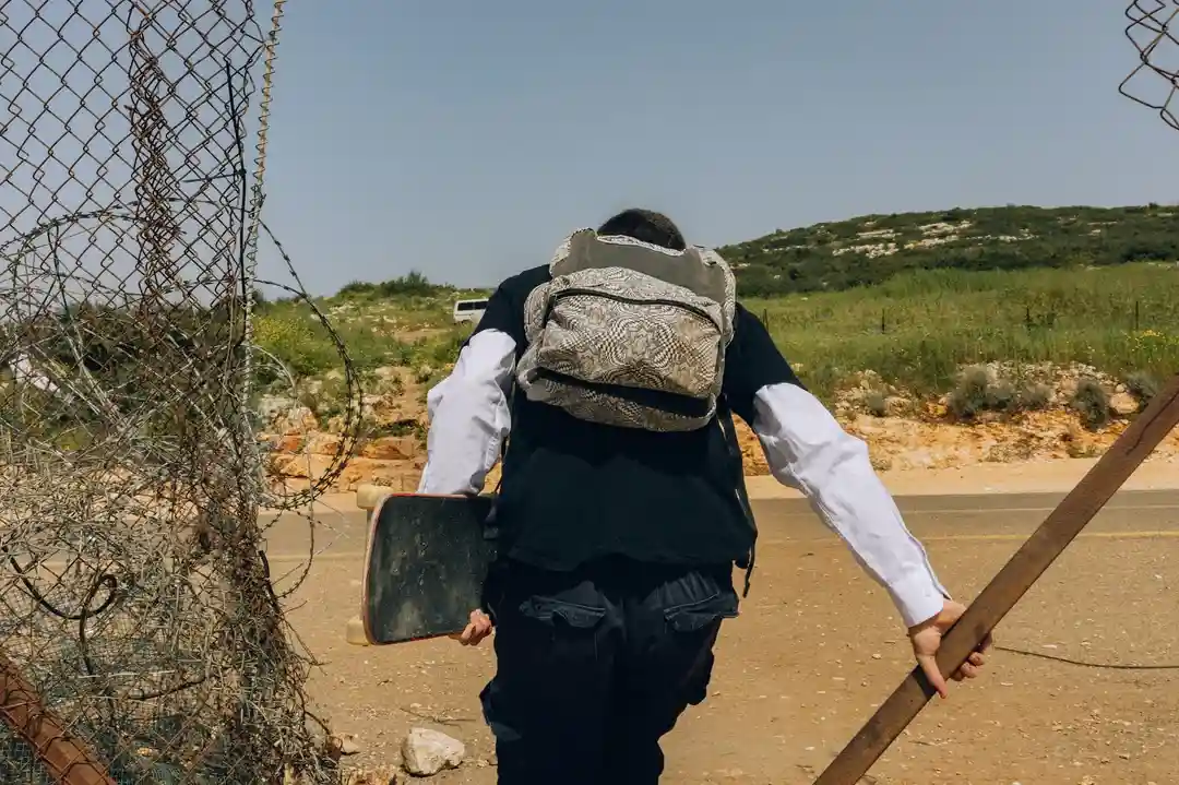 A person viewed from behind, wearing a black vest over a white shirt and carrying a patterned backpack, walks away from the…