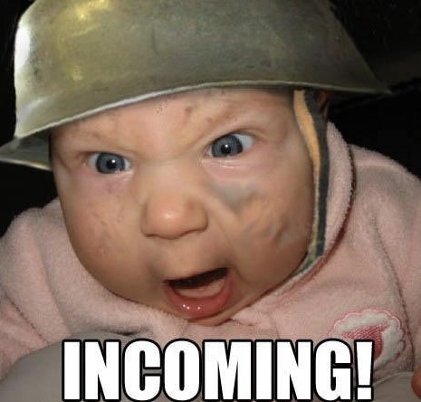 A close-up of a baby wearing a small, worn metal helmet, with wide, startled eyes and an open mouth, conveying shock or…