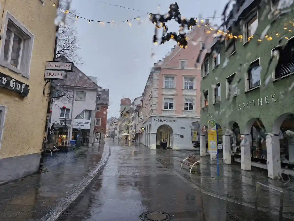 A rainy street view through a window with water droplets, showing European-style buildings including a hotel, pharmacy, and…