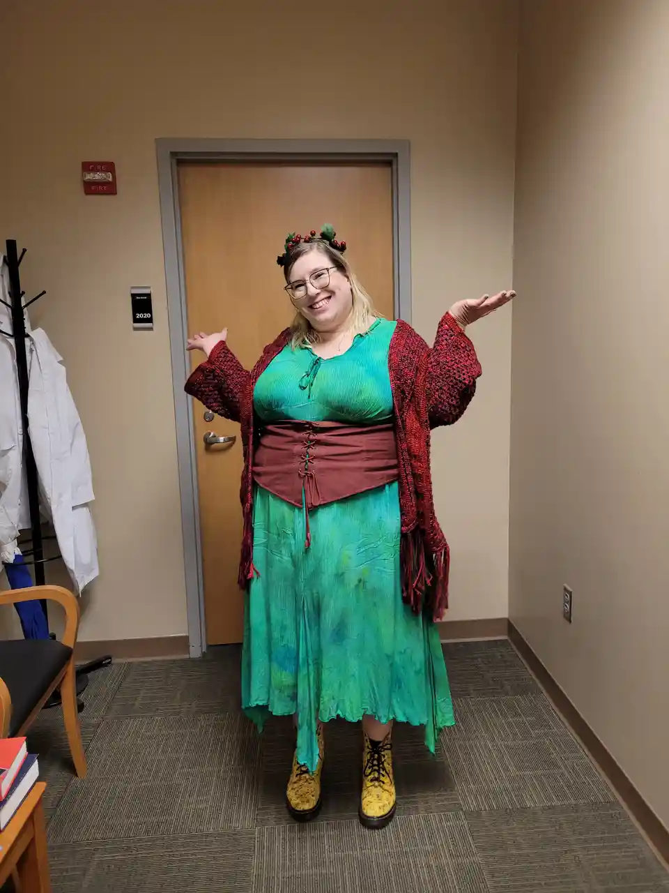 A smiling woman in a festive green dress, red crocheted cardigan, and a holly wreath stands in an office hallway in front of…