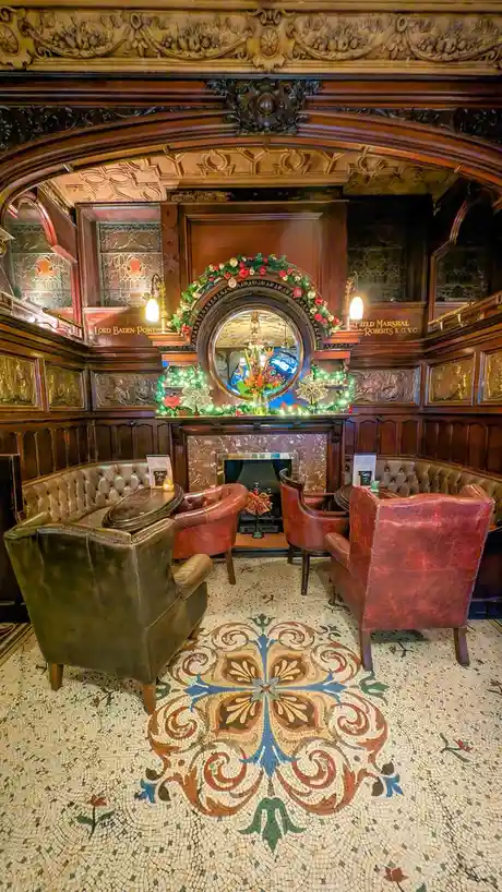 An ornate, wood-paneled room with a fireplace, red leather chairs, and a mosaic floor features a circular mirror above the…