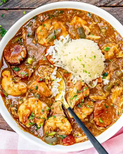 A top-down photo of a white bowl filled with a rich, brown stew containing shrimp, sausage slices, chicken, and vegetables…