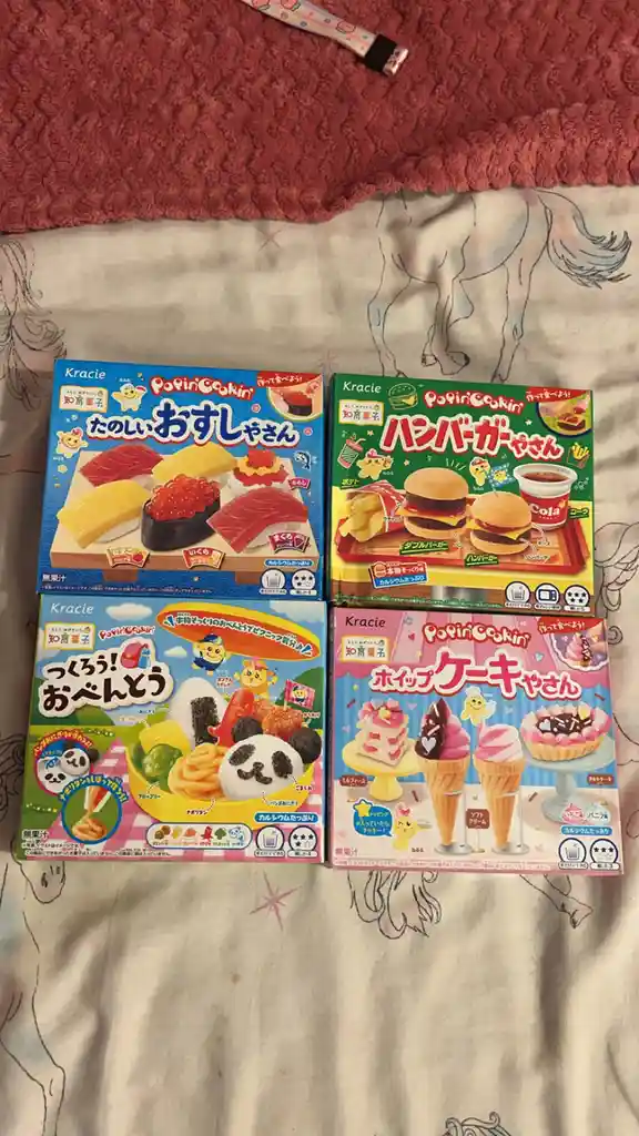 Four Kracie Popin' Cookin' Japanese edible craft kits are laid out on a bedspread with a horse pattern. Each box features…