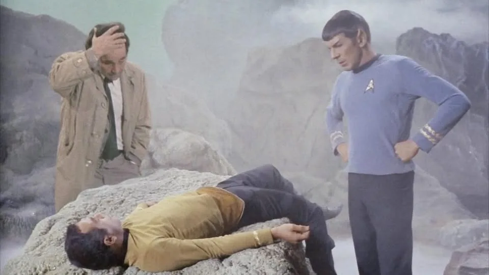 In a scene from Star Trek, Spock stands with hands on hips looking down at a fallen crewmate in a yellow uniform lying on a…