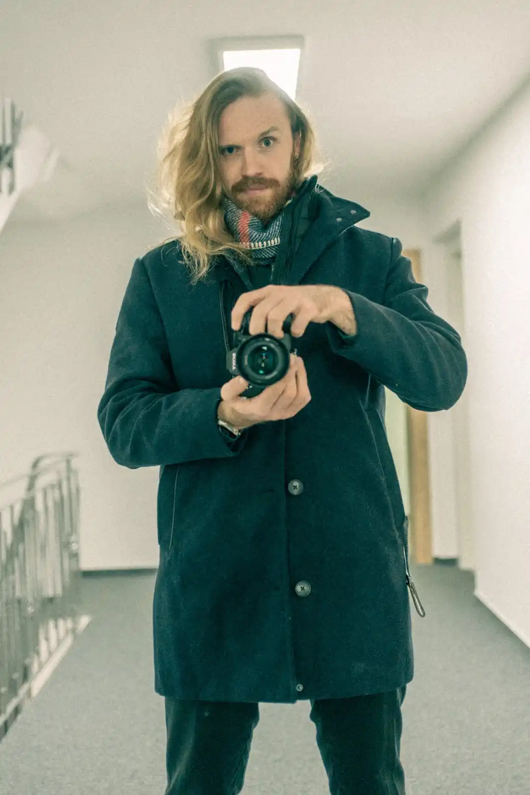 A man with long blonde hair and a beard, wearing a dark coat and scarf, holds a camera and looks directly at the viewer in a…