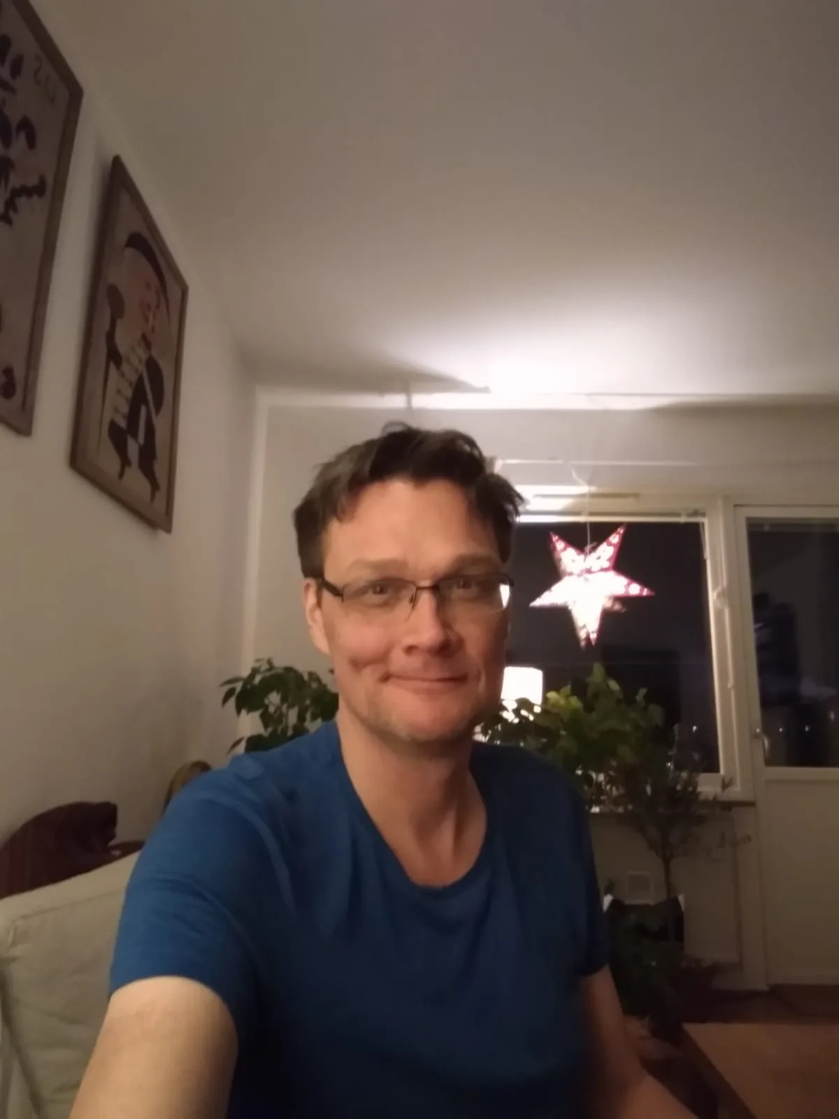 A man in a blue t-shirt and glasses smiles at the camera while sitting indoors, with a lit-up star decoration visible…